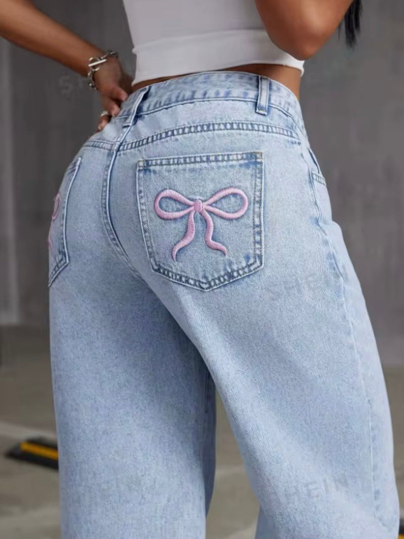 Close-up back view of a person wearing light-wash blue jeans with a pink bow embroidered on the back pocket, paired with a white cropped top.